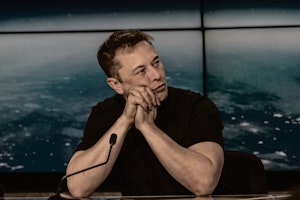 Elon Musk sitting at a table with a microphone looking bored