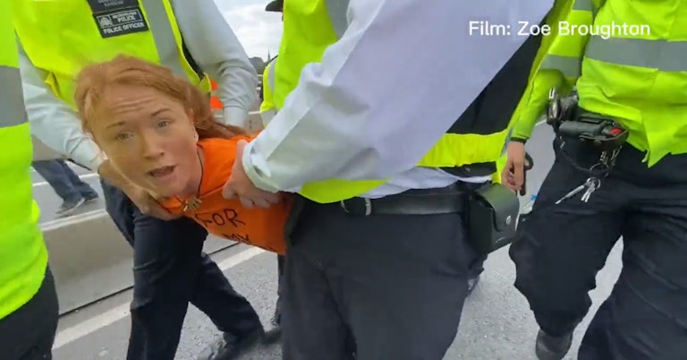 Young woman in an orange shirt speaking to the camera as a crowd of UK police officers carry her away by her arms and legs