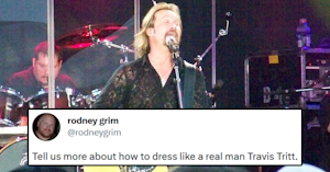 Travis Tritt singing and playing guitar on stage in a black blouse and silver necklace and tweet reading "Tell us more about how to dress like a real man Travis Tritt."