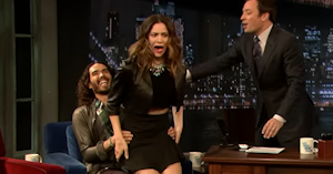 Katherine McPhee with her mouth open in shock as Russell Brand, seating in a chair behind her, smiles and grabs her by the hips, while Jimmy Fallon reaches for her.